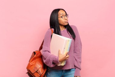Genç siyahi kadın omuz silkiyor, kafası karışık ve kararsız, kollarını kavuşturup şaşkın bakışlarla kuşkulanıyor. öğrenci konsepti
