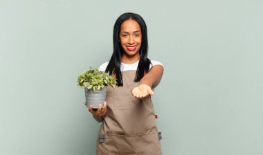 Genç siyahi kadın mutlu bir şekilde gülümsüyor, kendine güvenen, olumlu bakışlı, bir nesne ya da konsept sunuyor ve gösteriyor. Bahçıvan konsepti