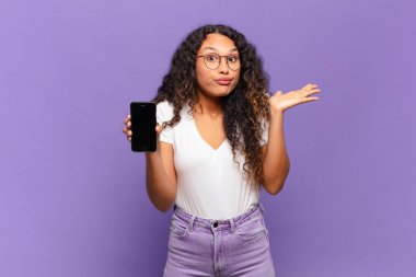 Genç İspanyol kadın şaşkın ve şaşkın hissediyor, şüpheci, farklı seçenekleri tartıyor ya da komik bir ifadeyle seçiyor. akıllı telefon kavramı