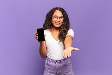 Genç İspanyol kadın mutlu bir şekilde gülümsüyor arkadaş canlısı, kendinden emin, olumlu bakışlı, bir nesne ya da konsept sunuyor ve gösteriyor. akıllı telefon kavramı