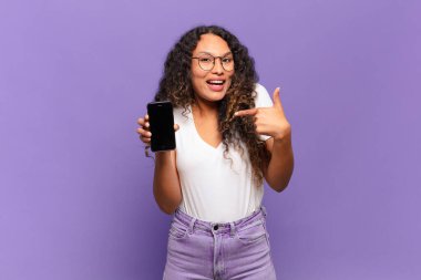 Genç İspanyol kadın mutlu, şaşkın ve gururlu hissediyor. Heyecanlı ve şaşkın bakışlarıyla kendini işaret ediyor. akıllı telefon kavramı