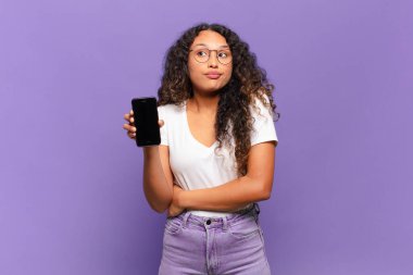 Genç İspanyol kadın omuz silkiyor, kafası karışık ve emin değil, kolları bağlı ve şaşkın görünüyor. akıllı telefon kavramı