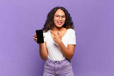 Genç İspanyol kadın heyecanlı ve şaşırmış görünüyor. Kenarlarını ve yukarıyı göstererek mekanı kopyalıyor. akıllı telefon kavramı