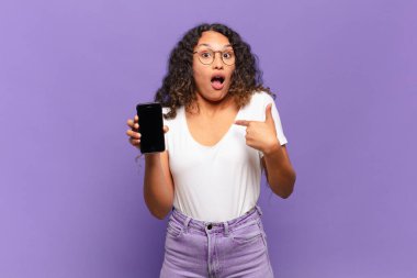 Genç İspanyol kadın şok olmuş ve ağzı açık, kendini işaret ederek şaşırmış görünüyor. akıllı telefon kavramı