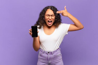 Genç İspanyol kadın mutsuz ve stresli görünüyor, intihar jesti elle silah işareti yapıyor, kafasını işaret ediyor. akıllı telefon kavramı