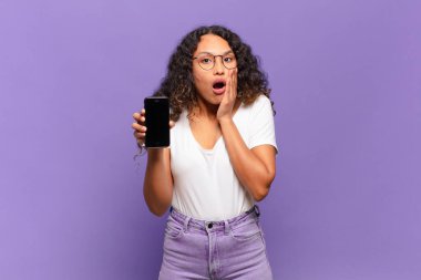 Genç İspanyol kadın şok olmuş ve korkmuş hissediyor, ağzı açık ve elleri yanaklarında korkmuş görünüyor. akıllı telefon kavramı