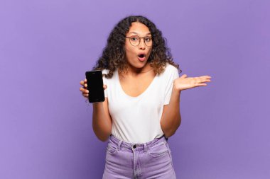 Genç İspanyol kadın şaşırmış ve şok olmuş görünüyor. Çenesi düşmüş ve eli açık bir objeyi tutuyor. akıllı telefon kavramı