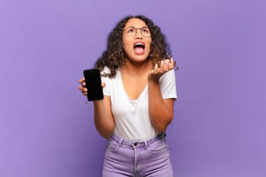 Genç İspanyol kadın umutsuz ve sinirli görünüyor, stresli, mutsuz ve sinirli, bağırıyor ve çığlık atıyor. akıllı telefon kavramı