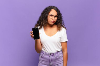 young hispanic woman feeling puzzled and confused, with a dumb, stunned expression looking at something unexpected. smart phone concept