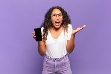 Genç İspanyol kadın aşırı derecede şok olmuş ve şaşırmış, endişeli ve paniklemiş hissediyor, stresli ve dehşete kapılmış bir bakışla. akıllı telefon kavramı