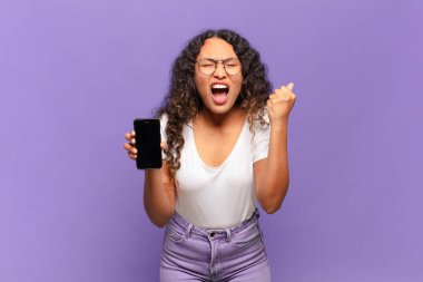 Genç İspanyol kadın öfkeli bir ifadeyle agresif bir şekilde bağırıyor ya da yumruklarıyla başarıyı kutluyor. akıllı telefon kavramı