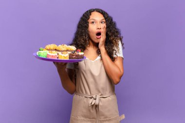 Genç İspanyol kadın şok olmuş ve korkmuş hissediyor, ağzı açık ve elleri yanaklarında korkmuş görünüyor. kek pişirme kavramı