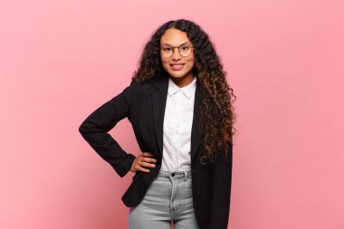 Genç İspanyol kadın mutlu bir şekilde gülümsüyor, eli kalçasında ve kendinden emin, olumlu, gururlu ve arkadaş canlısı bir tavırla. iş kavramı