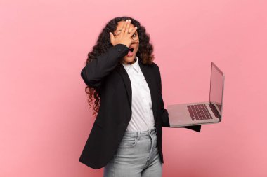 Genç İspanyol kadın şok olmuş, korkmuş ya da korkmuş görünüyor. Yüzü elle kaplanmış ve parmaklarının arasından gizlice bakıyor. dizüstü bilgisayar