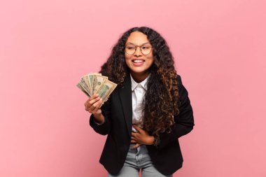 Genç İspanyol kadın komik bir şakaya yüksek sesle gülüyor, mutlu ve neşeli hissediyor, eğleniyor. Dolar banknotları konsepti