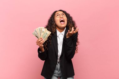 Genç İspanyol kadın umutsuz ve sinirli görünüyor, stresli, mutsuz ve sinirli, bağırıyor ve çığlık atıyor. Dolar banknotları konsepti