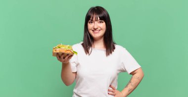 Mutlu bir şekilde gülümsüyor, eli kalçasında ve kendinden emin, olumlu, gururlu ve arkadaş canlısı bir tavırla.