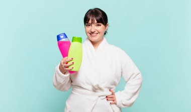 Bornoz giyen genç bir kadın mutlu bir şekilde gülümsüyor elleri kalçasında ve kendinden emin, olumlu, gururlu ve arkadaş canlısı bir tavırla.