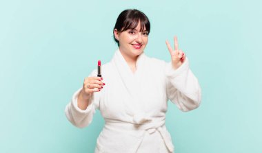 Bornoz giymiş genç bir kadın gülümsüyor ve arkadaş canlısı görünüyor, iki ya da iki numarayı gösteriyor, eli önde, geriye doğru sayıyor.