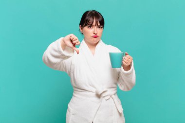 Bornoz giyen genç bir kadın kızgın, sinirli, hayal kırıklığına uğramış ya da hoşnutsuz, ciddi bir bakışla başparmağını kaldırıyor.