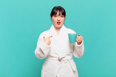 Bornoz giymiş genç bir kadın şok olmuş ve ağzı açık bir şekilde kendini işaret ederek şaşırmış görünüyor.