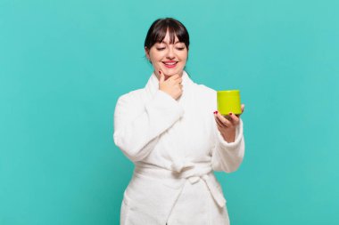 Bornoz giyen genç bir kadın mutlu, kendinden emin bir ifadeyle gülümsüyor, çenesinde elleri, merak ediyor ve yan tarafa bakıyor.