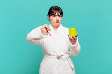 Bornoz giyen genç bir kadın kızgın, sinirli, hayal kırıklığına uğramış ya da hoşnutsuz, ciddi bir bakışla başparmağını kaldırıyor.