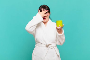 Bornoz giymiş genç bir kadın şok olmuş, korkmuş ya da korkmuş görünüyor. Yüzü elle kaplanmış ve parmaklarının arasından gizlice bakıyor.