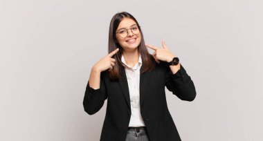 Genç iş kadını kendinden emin bir şekilde gülümsüyor. Geniş bir gülümseme, pozitif, rahat, tatmin olmuş bir tavır sergiliyor.