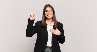 Genç iş kadını mutlu, kendinden emin ve güvenilir görünüyor, gülümsüyor ve olumlu bir tavırla zafer işareti gösteriyor.
