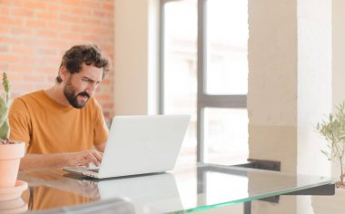 Evde dizüstü bilgisayarla çalışan sakallı genç bir adam.