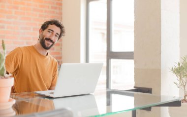 Genç sakallı adam mutlu bir şekilde gülümsüyor, eli kalçasında ve kendinden emin, pozitif, gururlu ve arkadaş canlısı bir tavırla ve bir dizüstü bilgisayarla çalışıyor.