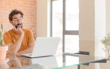 Genç sakallı adam mutlu bir şekilde gülümsüyor ve hayal kuruyor ya da şüphe ediyor, yan tarafa bakıyor ve bir dizüstü bilgisayarla çalışıyor.