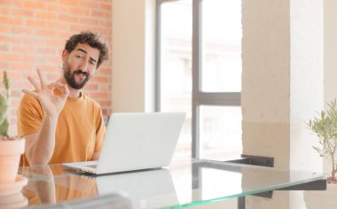 Genç sakallı adam mutlu, rahat ve tatmin olmuş hissediyor. İyi bir jest yapıyor, gülümsüyor ve dizüstü bilgisayarla çalışıyor.