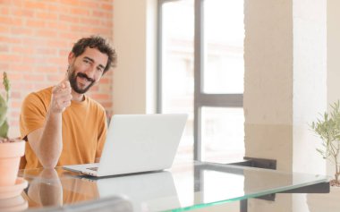 Genç sakallı adam kameraya tatmin olmuş, kendinden emin, dostane bir gülümsemeyle bakıyor, seni seçiyor ve bir dizüstü bilgisayarla çalışıyor.