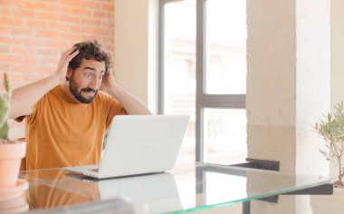 Genç sakallı adam kendini stresli, endişeli, endişeli ya da korkmuş hissediyor. Elleri başının üstünde. Yanlışlıkla panikliyor ve dizüstü bilgisayarla çalışıyor.