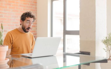 Genç sakallı adam şok olmuş ya da şaşırmış görünüyor, ağzı açık bakıyor ve vay canına diyor ve bir dizüstü bilgisayarla çalışıyor.