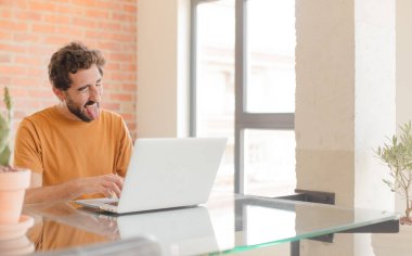 Genç sakallı, neşeli, kaygısız, asi tavırlı, şakacı ve dilsiz, eğlenen ve dizüstü bilgisayarla çalışan bir adam.