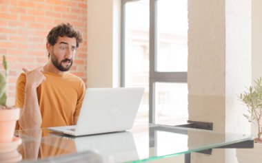 Genç sakallı bir adam stresli, endişeli, yorgun ve hayal kırıklığına uğramış, gömlek yakalı, sorunlu görünüyor ve bir dizüstü bilgisayarla çalışıyor.