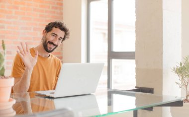 Genç sakallı adam mutlu ve neşeyle gülümsüyor, el sallıyor, seni selamlıyor, ya da hoşça kal diyor ve bir dizüstü bilgisayarla çalışıyor.