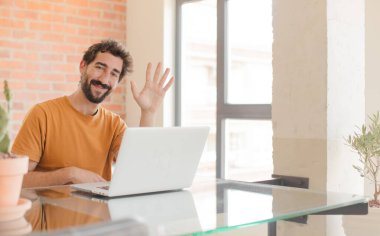 Genç sakallı adam gülümsüyor ve arkadaş canlısı görünüyor, 5 ya da 5 numarayı gösteriyor, geriye doğru sayıyor ve dizüstü bilgisayarla çalışıyor.
