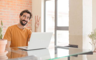 Genç sakallı adam gülümsüyor ve arkadaş canlısı görünüyor. Elleri önde, geriye doğru sayıyor ve dizüstü bilgisayarla çalışıyor.