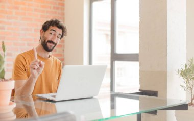 Genç sakallı adam gururla gülümsüyor ve gururla bir numaralı pozu veriyor. Kendini lider gibi hissediyor ve dizüstü bilgisayarla çalışıyor.