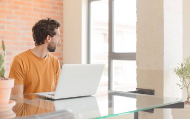 Profil görünümlü genç sakallı adam ileriyi kopyalamaya çalışıyor, düşünüyor, hayal ediyor ya da hayal kuruyor ve bir dizüstü bilgisayarla çalışıyor.