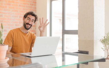 Genç sakallı adam gülümsüyor ve arkadaş canlısı görünüyor. Elleri önde, geriye doğru sayıyor ve dizüstü bilgisayarla çalışıyor.