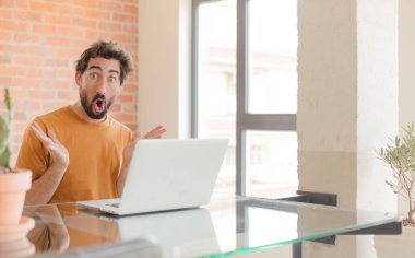 Genç sakallı adam, ağzı açık ve şaşkın, şok olmuş ve hayretler içinde inanılmaz bir sürprizle ve bir dizüstü bilgisayarla çalışıyor.