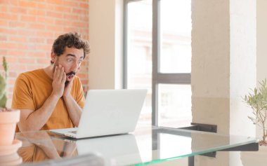 Genç sakallı adam şok olmuş ve korkmuş hissediyor, ağzı açık ve elleri yanaklarında korkmuş görünüyor ve dizüstü bilgisayarla çalışıyor.