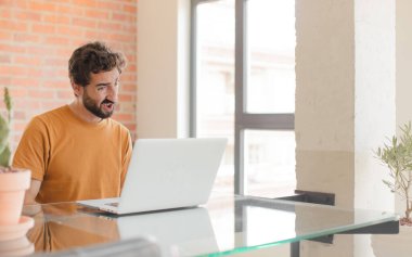 young bearded man feeling puzzled and confused, with a dumb, stunned expression looking at something unexpected and working with a laptop