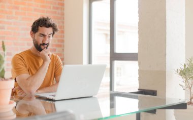 Genç sakallı adam düşünüyor, kuşkulu ve şaşkın hissediyor, farklı seçeneklerle, hangi kararı vereceğini merak ediyor ve bir dizüstü bilgisayarla çalışıyor.