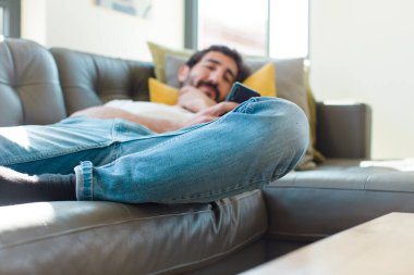 Genç sakallı adam akıllı telefonuyla kanepede dinleniyor.